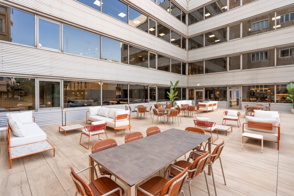 Amplia terraza exterior estilo chill-out en edificio de oficinas o coworking, con mesas, sillas, sofás modernos y plantas, ideal para descanso, eventos o trabajo al aire libre.