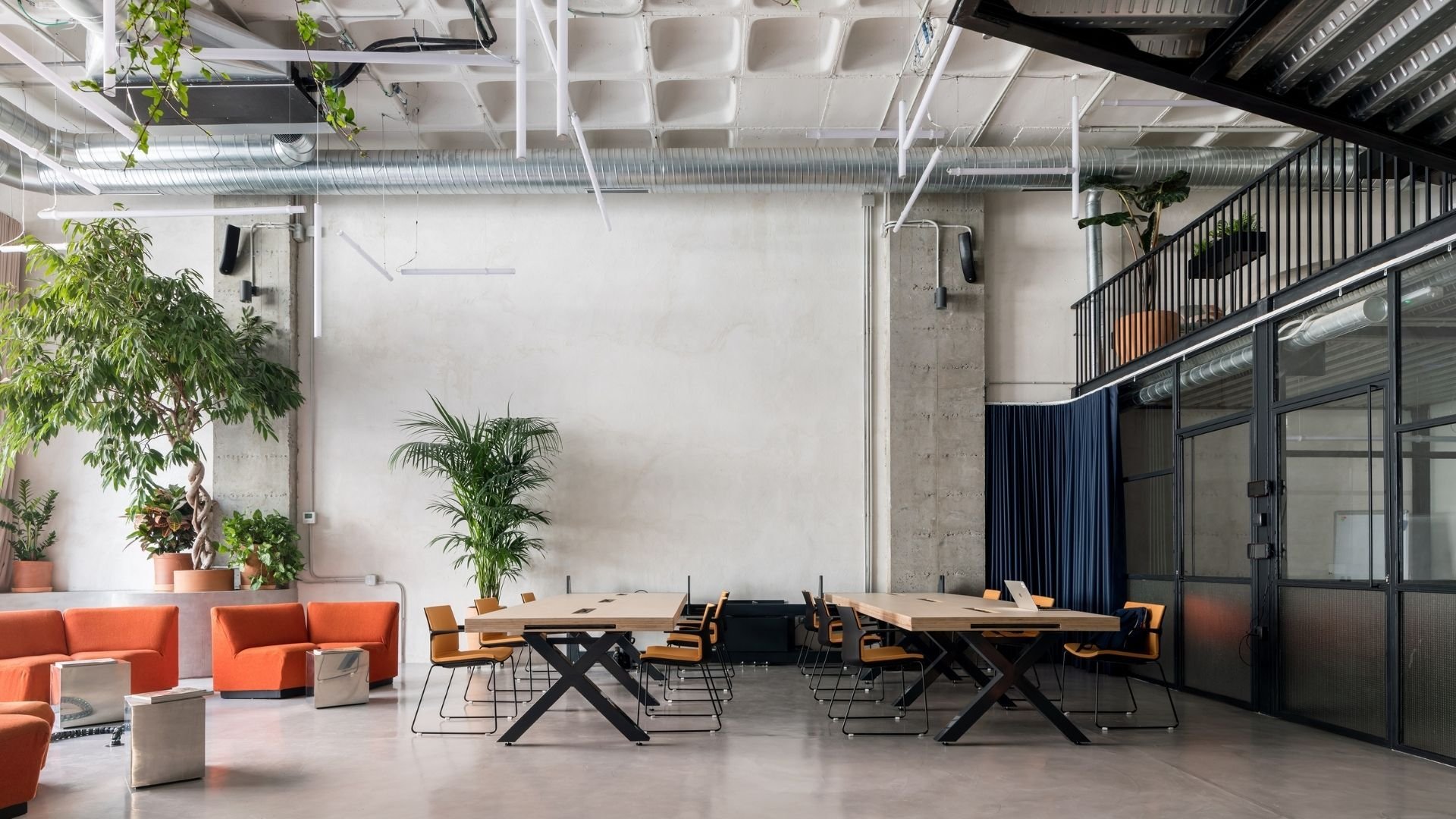 Espacio de oficina híbrida moderna con techos altos industriales, mesas de trabajo compartido, abundante vegetación y zona lounge con sofás naranjas para colaboración informal.